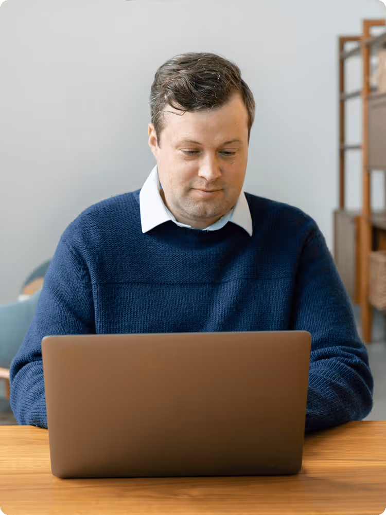 Man in a blue sweater working on a laptop at a wooden table indoors.
