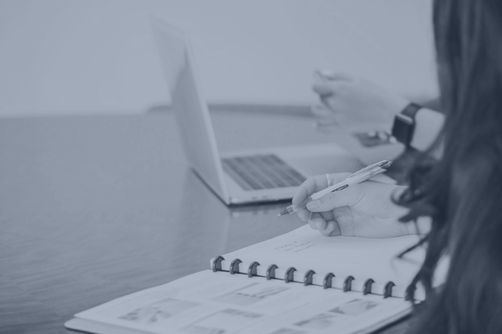 A person writing notes in a notebook beside a laptop